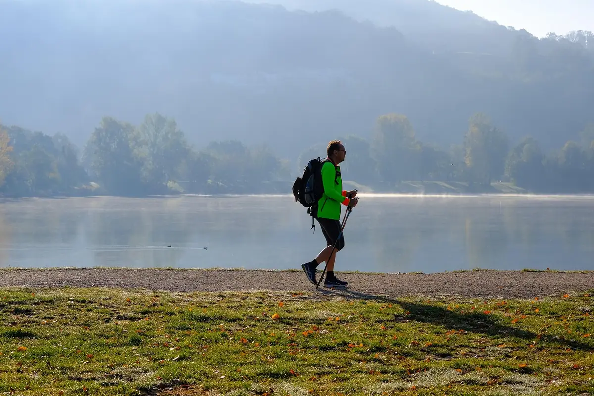 Mężczyzna z plecakiem maszeruje z kijkami do nordic walking po trawie nad jeziorem, w tle zalesione wzgórza