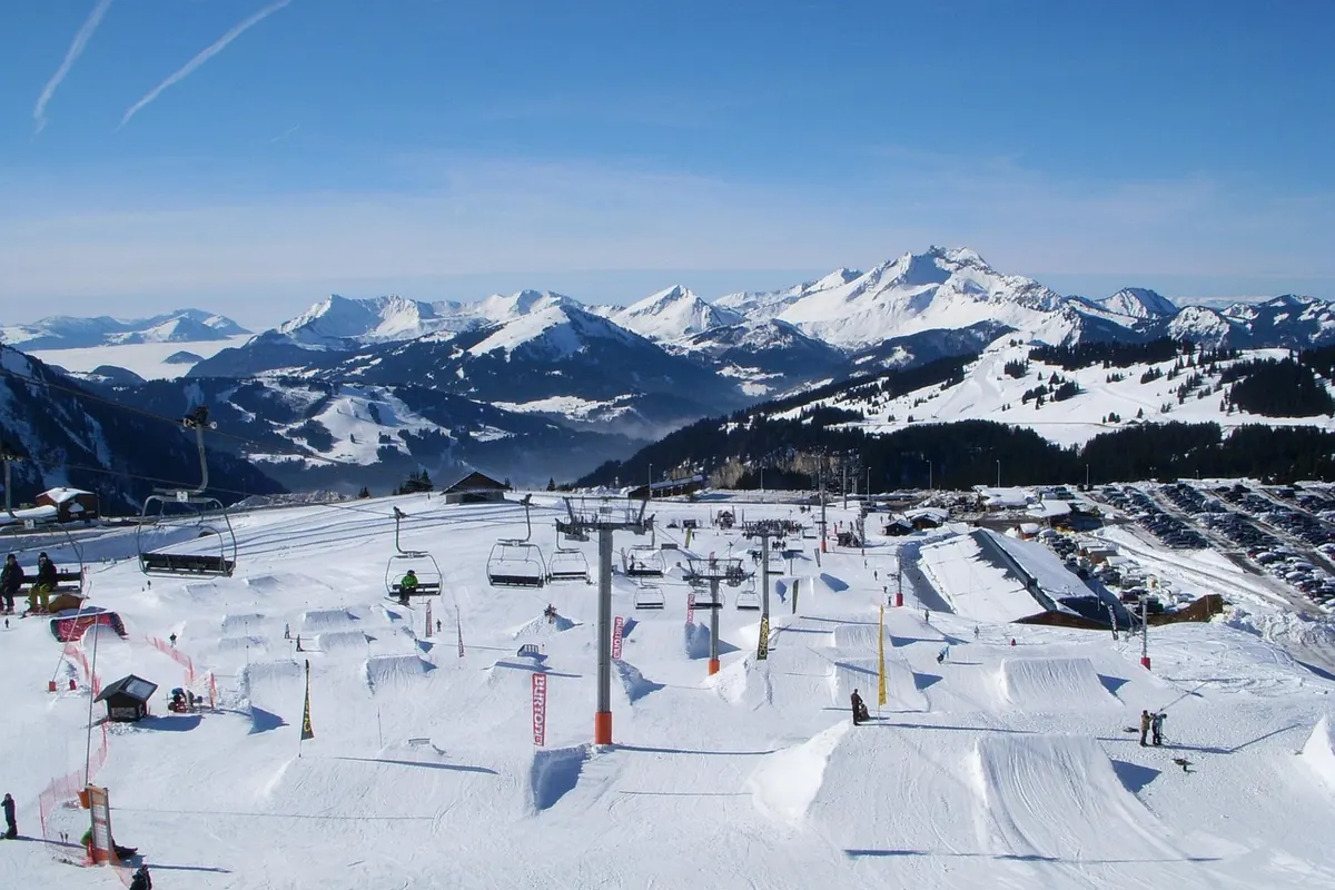 Snowpark na szerokim stoku z rampami i przeszkodami, obok wyciąg, w tle pasmo ośnieżonych gór