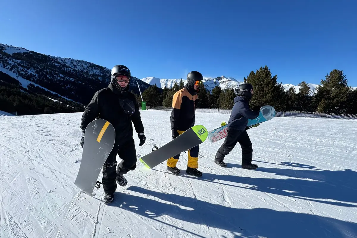 Trzy osoby w kaskach idą po płaskim śniegu z deskami snowboardowymi w rękach