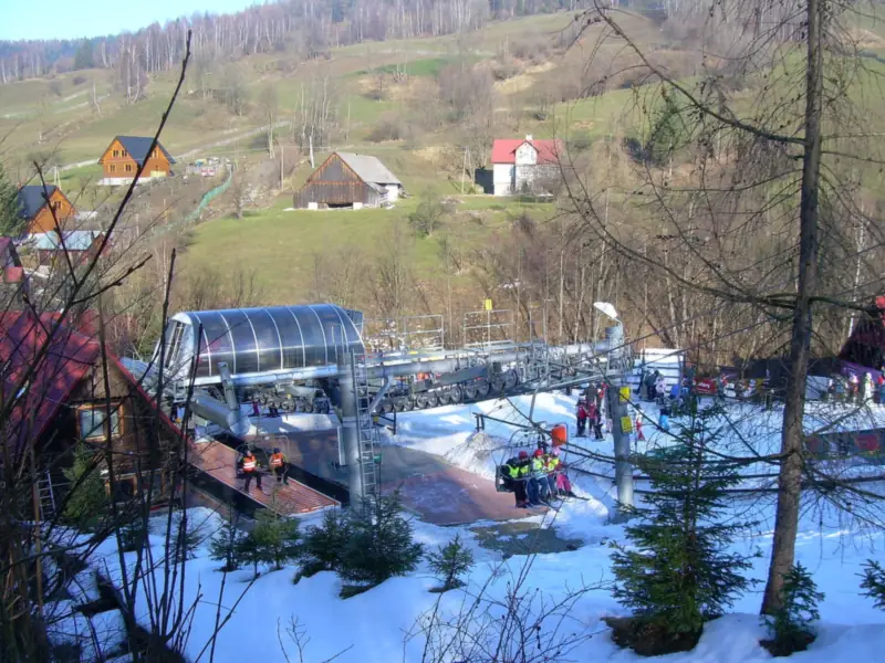 Dolna stacja kolei krzesełkowej w Wierchomli z narciarzami na ośnieżonym placu, w tle zbocze z domami