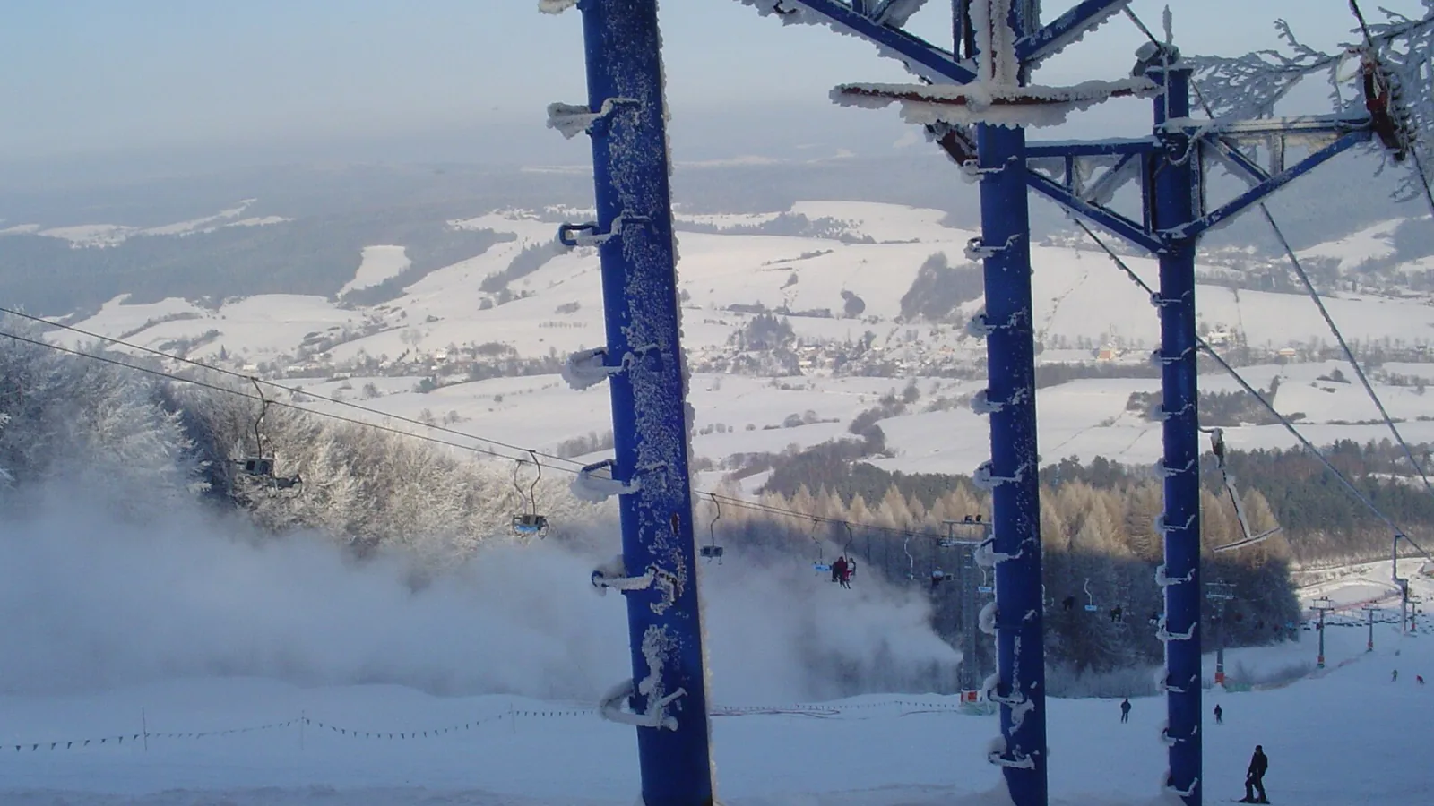Niebieskie podpory wyciągu nad ośnieżoną trasą Laworty, w dole zaśnieżone pola i las