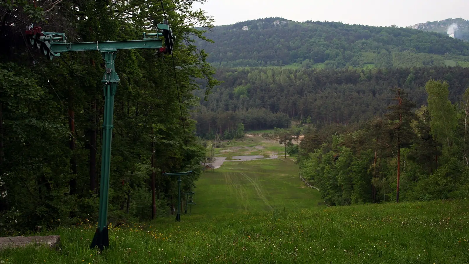 Słup wyciągu orczykowego przy zielonym stoku w Morsku, w tle zalesione wzgórza Jury latem