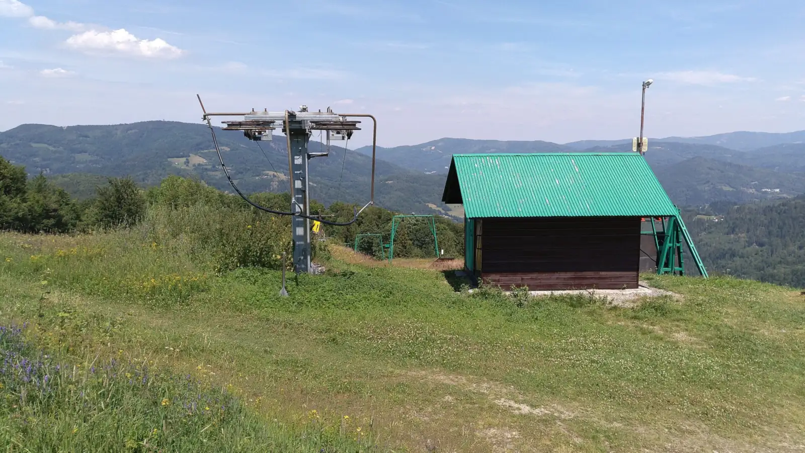Wyciąg orczykowy na trawiastym zboczu Soszowa z drewnianą budką, w tle zalesione góry latem