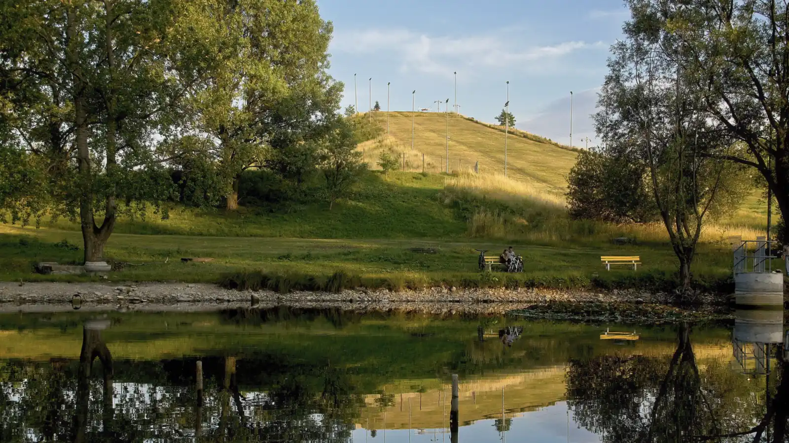 Trawiaste wzgórze stoku narciarskiego Środula odbite w stawie Florek, wokół drzewa parku latem