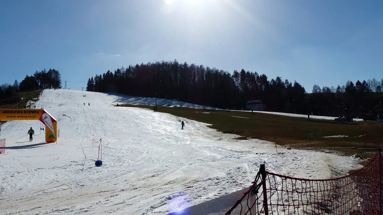 Trasa zjazdowa w Koszałkowie w słońcu pod koniec sezonu, narciarze na stoku i las na grzbiecie