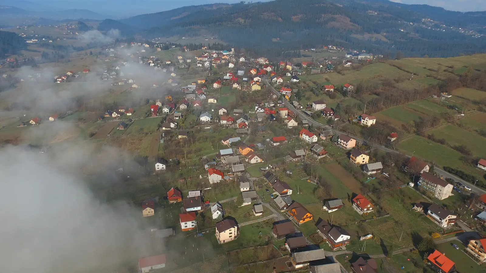 Widok z góry na zabudowę Istebnej wśród pól, nad doliną mgła i zalesione grzbiety