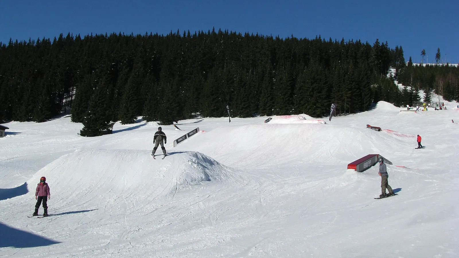 Snowpark z hopkami i poręczami na ośnieżonej polanie wśród świerków, narciarze i snowboardziści