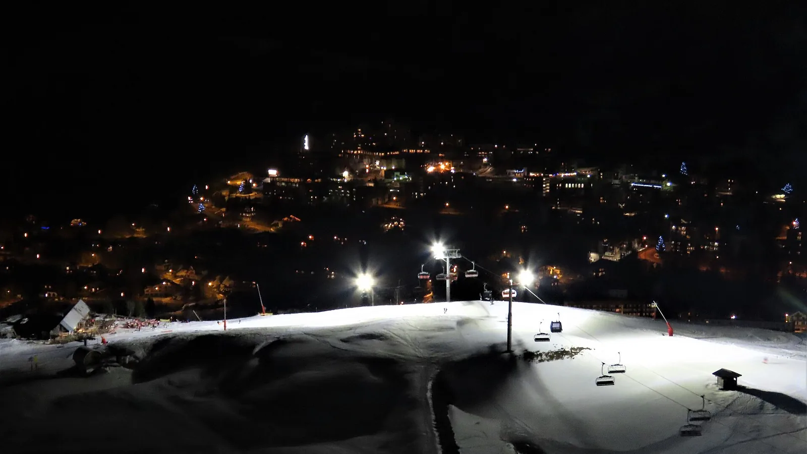 Oświetlona nocą trasa narciarska nad rozświetloną miejscowością Les Deux Alpes w dolinie