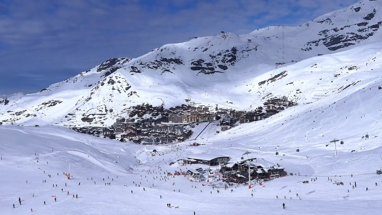 Szeroki ośnieżony stok z narciarzami prowadzący do zabudowy Val Thorens u stóp zaśnieżonych grzbietów